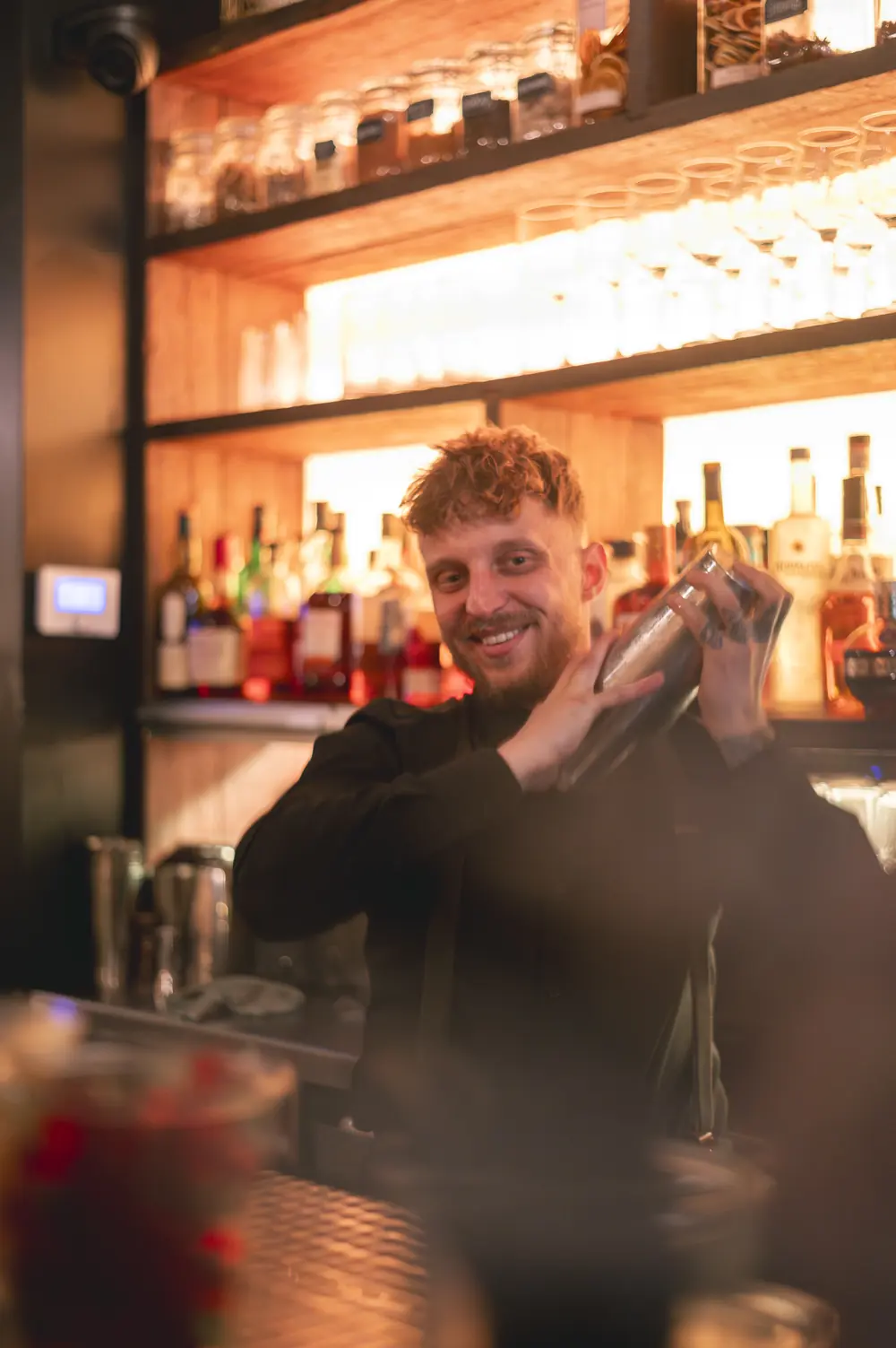 Bartender van WonderBar Genk met cocktailshaker, lachend voor de gevulde flessenkast