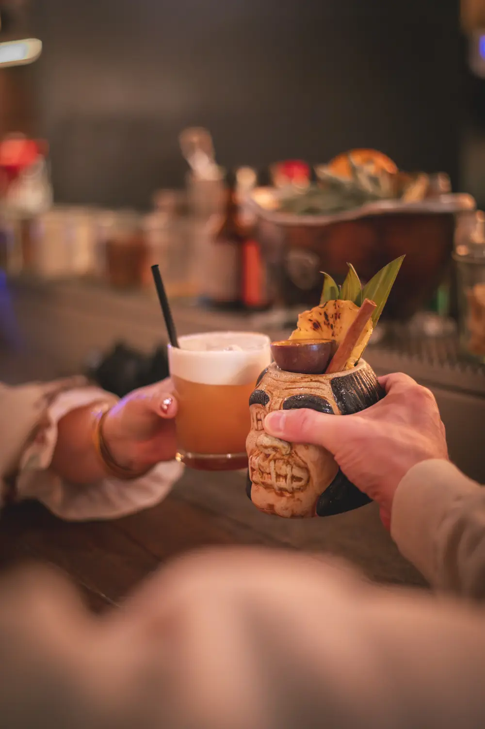 Twee gasten klinken hun glazen samen aan de bar van WonderBar Genk, een whisky sour en een tiki-mok met ananas-garnish en kaneelstok