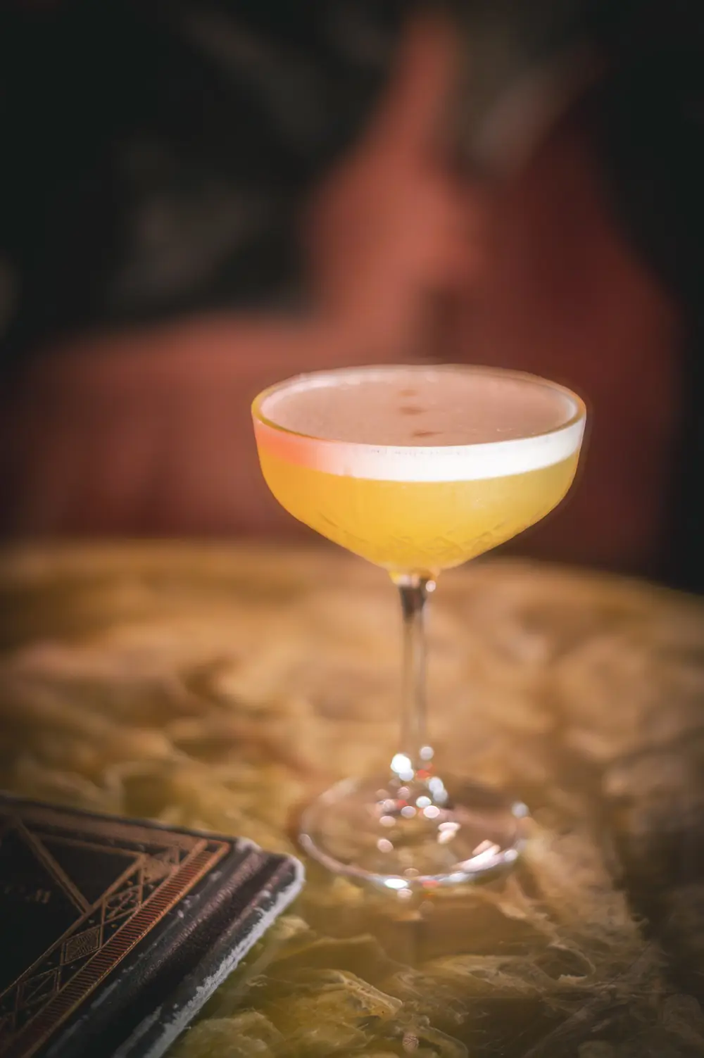 Coupe-glas met whisky sour-stijl cocktail op gouden marmeren tafel naast antiek receptenboek in WonderBar Genk