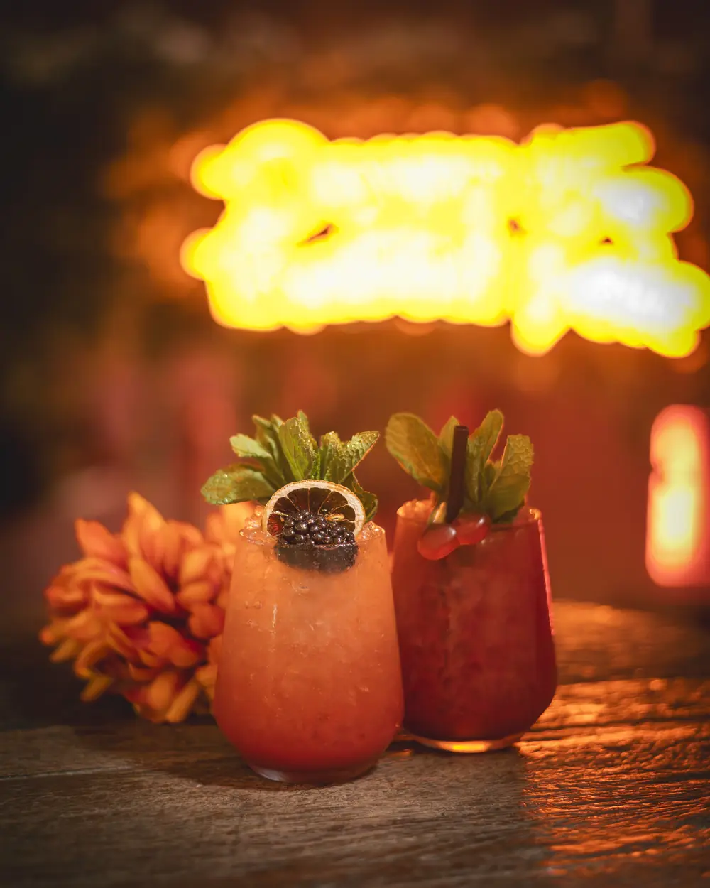 Twee cocktails op een tafel onder de oranje neon &lsquo;Down the Rabbit hole&rsquo; in WonderBar Genk, gegarneerd met mint, blackberry en citrusschijf