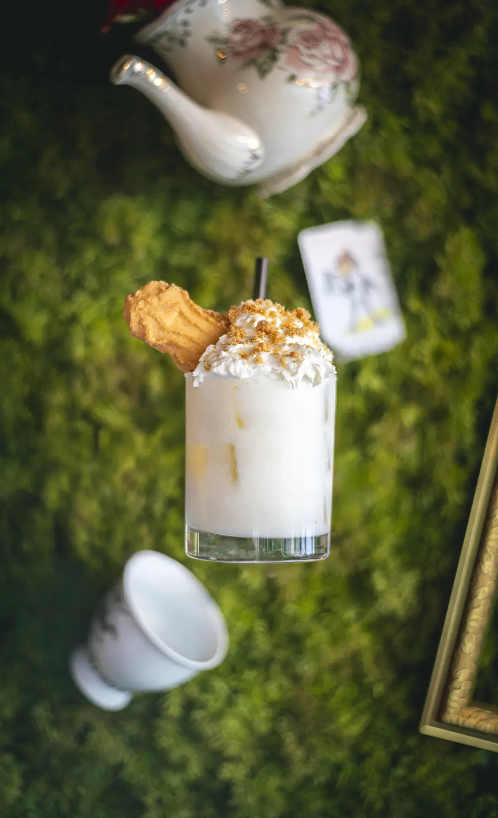 Romige cocktail met koekje en crumble op een mosgroene ondergrond, met bloemig theepotje, omgevallen theekopje, speelkaart en gouden lijst eromheen in WonderBar Genk